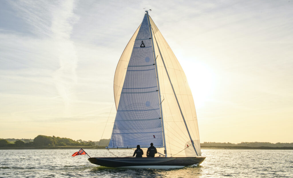 Large yacht with main sail and jib up sailing though inshore water at sunrise with two passengers onboard.