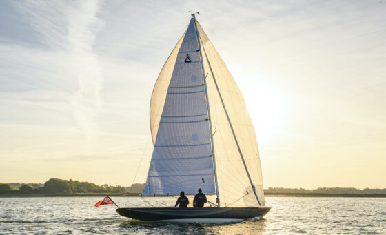 Large yacht with main sail and jib up sailing though inshore water at sunrise with two passengers onboard.
