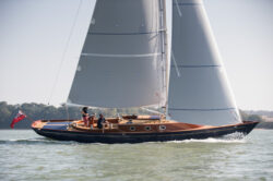 Large yacht with main sail and jib up sailing though inshore water with two passengers onboard.