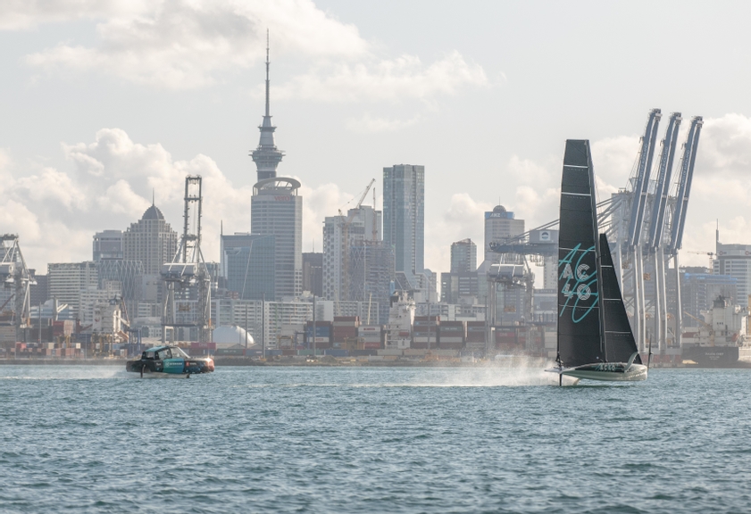 AC40 America's Cup boat foiling at speed through the water with hydrogen powered chase boat foiling behind.