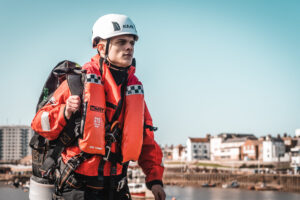 Hombre vestido con equipo de seguridad marina al lado de un puerto con el agua y la ciudad al fondo.