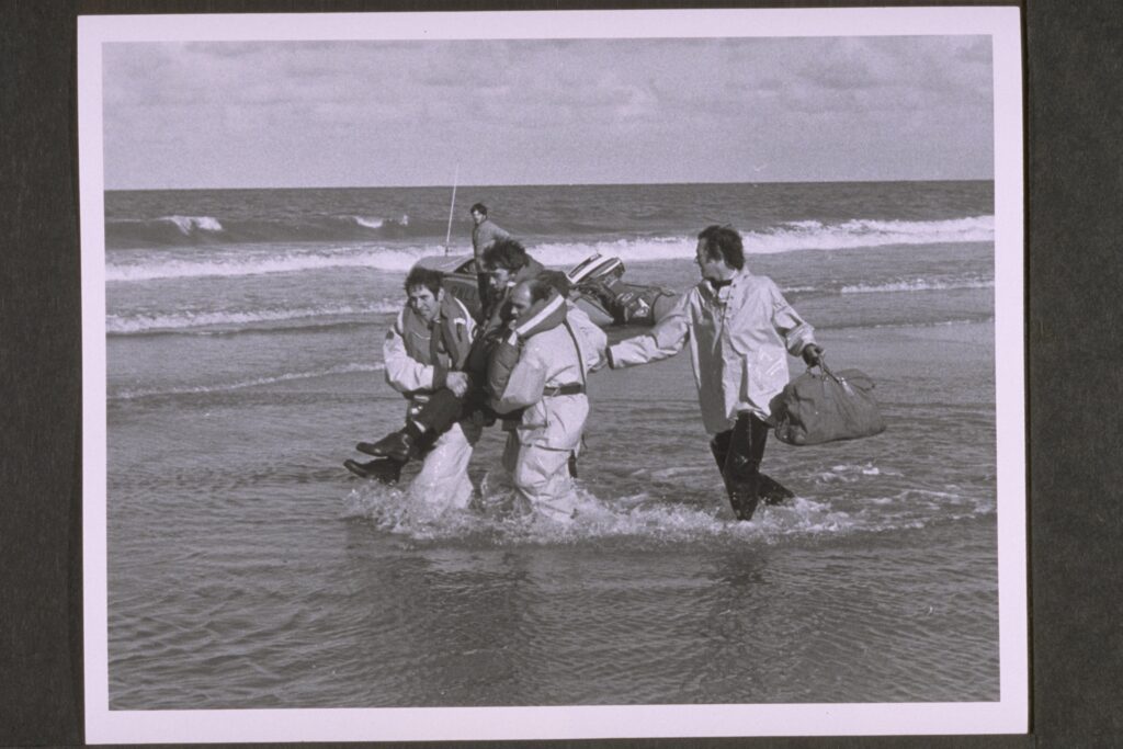 克罗默。 D级救生艇在海滩上。 两名救生艇船员抬着一名男子，另一名从后面扶着他。 船员从左到右 Ted Luckin（舵手）、Eric Love 和 Chris Craske。