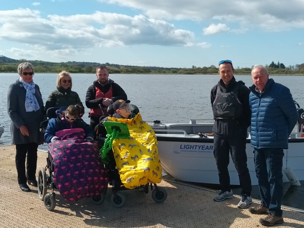 Membros do Wheely Boat Trust com o barco acessível para cadeira de rodas nas margens do lago