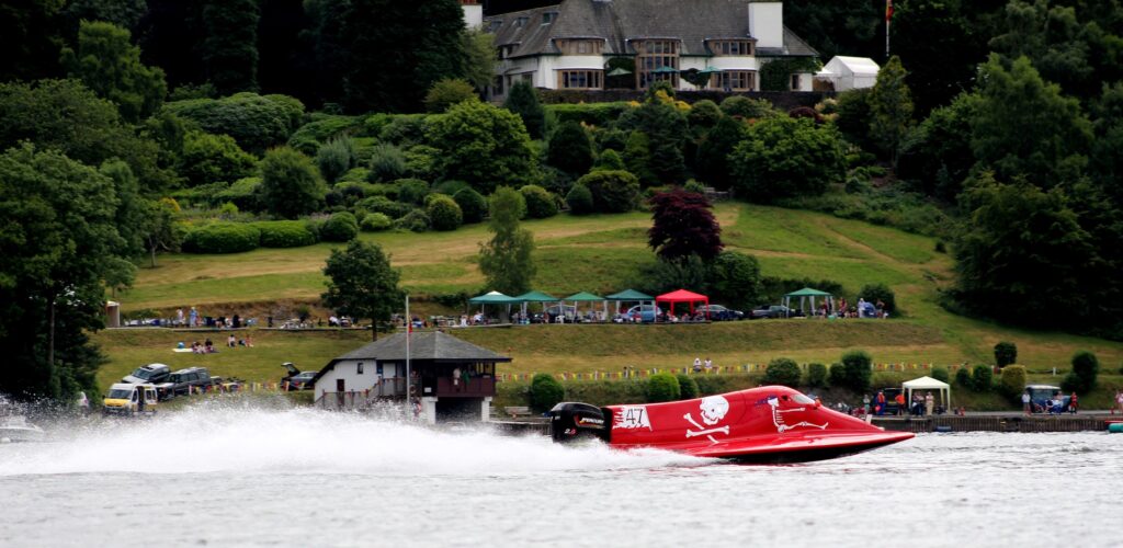 A red power boat blasts up a lake with cars and spectators in the background