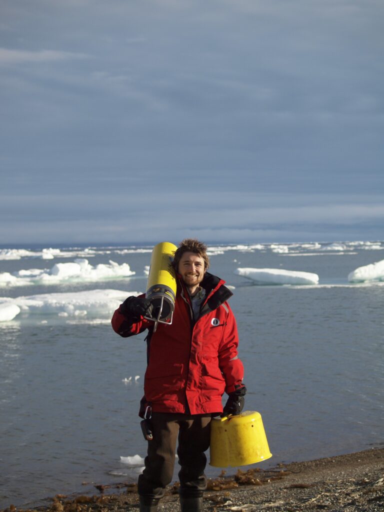 Bearded man on beach in red jacket carrying yellow  research gear. The waves crash behind him.
