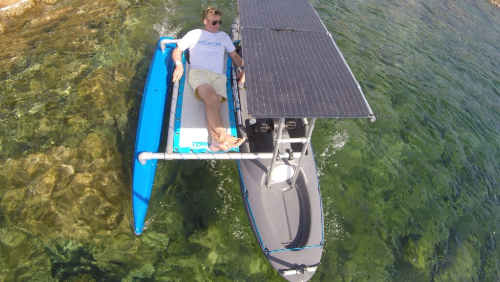 رجل يتكئ على امتداد منصة بيداياك في وسط الماء. توفر اللوحة الشمسية العلوية الظل على هذا الكاياك الذي يعمل بالطاقة الشمسية