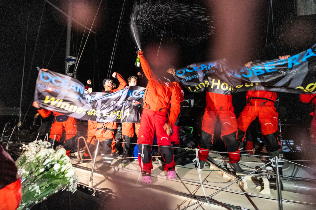 Crew of yacht celebrate by standing on deck with a banner as skipper - wearing pink crocs - raises a bottle spraying champagne above her head in triumph