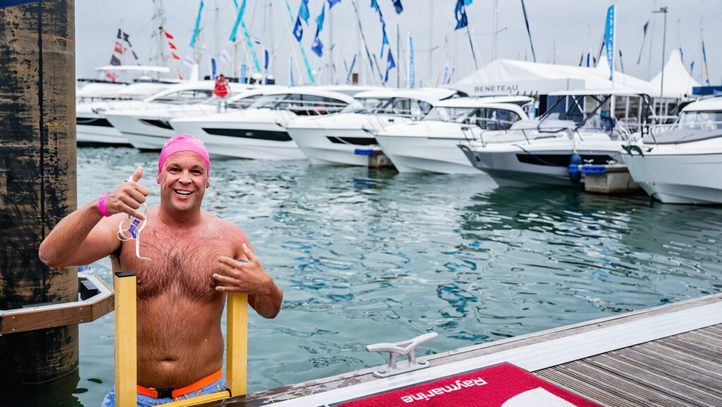 El nadador sale del agua por una escalera amarilla; Lleva un sombrero rosa y hay un montón de barcos al fondo.