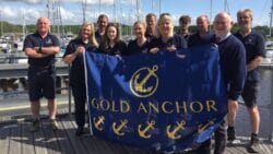 The Buckler's Hard Yacht Harbour team stand on a pontoon and hold up a flag with gold anchors on it (five gold anchors)