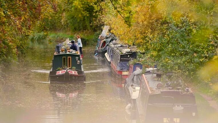 A canal boat meanders along a canal past several other boars moored to banks