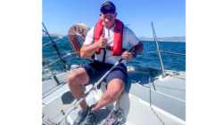 Een zeeman steekt vrolijk zijn duim omhoog vanaf de achtersteven van een boot, terwijl de zee zich in de verte uitstrekt