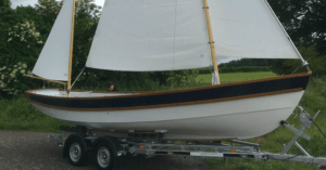 The hull of a boat on a trailer with sails up. It's a Drascombe Gig as ordered by the French Navy