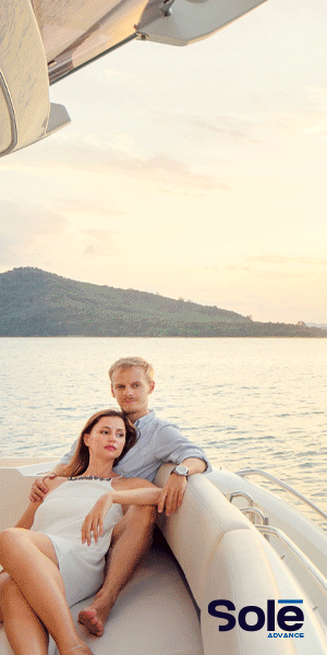 A couple relaxing on a yacht at sunset with a scenic view of a hill in the background and the logo "Solé" in the corner.