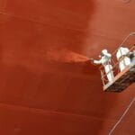 workers in white suits spray ship hull with red coating