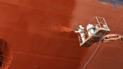 workers in white suits spray ship hull with red coating