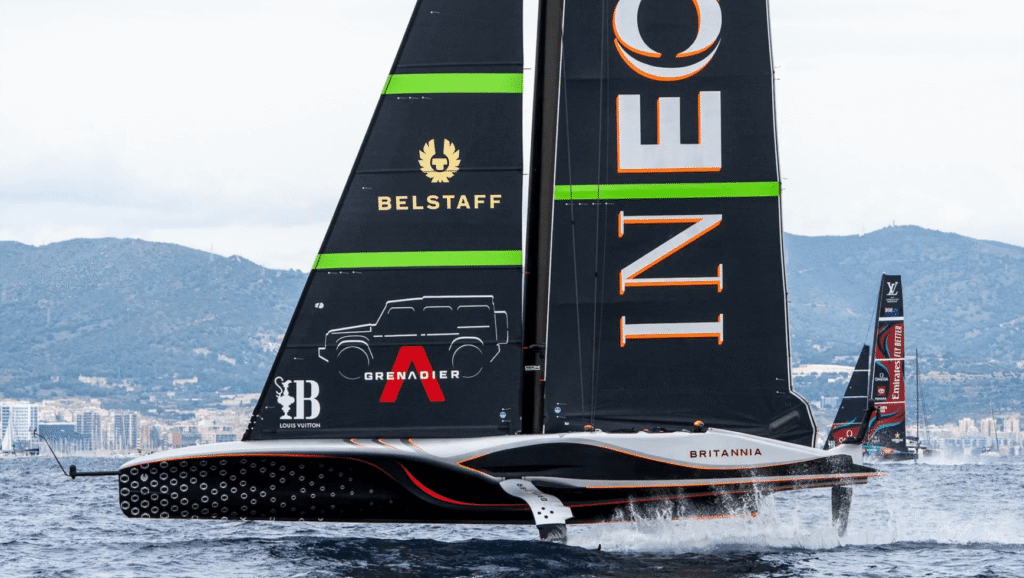 A sleek black sailboat with the words INEOS and BRITANNIA on its sails glides over the water, echoing the precision of New Zealand Rugby. The boat features logos for Belstaff and Grenadier, with green and red accents. Mountains and cityscape are visible in the background under a cloudy sky. marine industry news