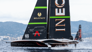 A sleek black sailboat with the words INEOS and BRITANNIA on its sails glides over the water, echoing the precision of New Zealand Rugby. The boat features logos for Belstaff and Grenadier, with green and red accents. Mountains and cityscape are visible in the background under a cloudy sky. marine industry news
