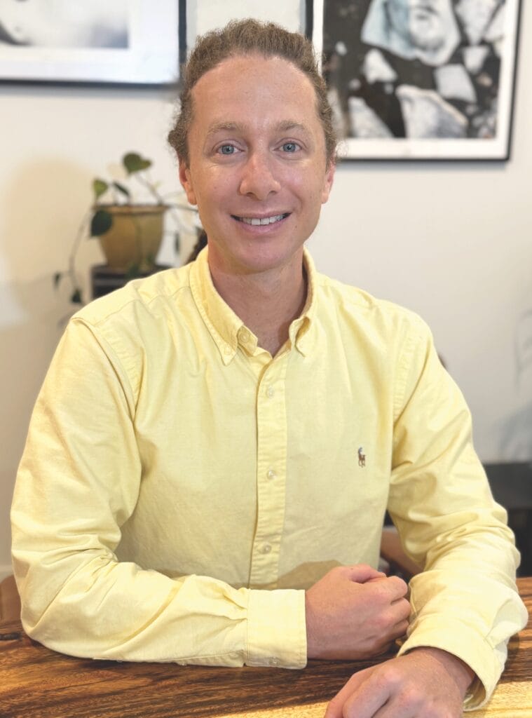 Marcus Hamilton wears a yellow shirt. He sits behind a desk and smiles at the camera as he talks about antifoul trends for 2025