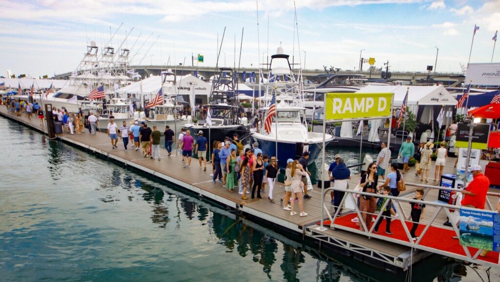 People stroll along a bustling marina dock lined with boats, many displaying American flags, during the Miami Boat Show 2025. A prominent RAMP D sign stands out as tents and vendors bustle in the background under a partly cloudy sky. marine industry news