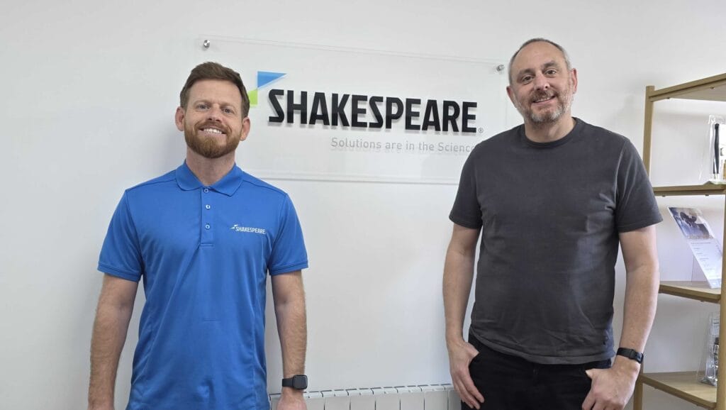Two men standing by Shakespeare Marine logo in office