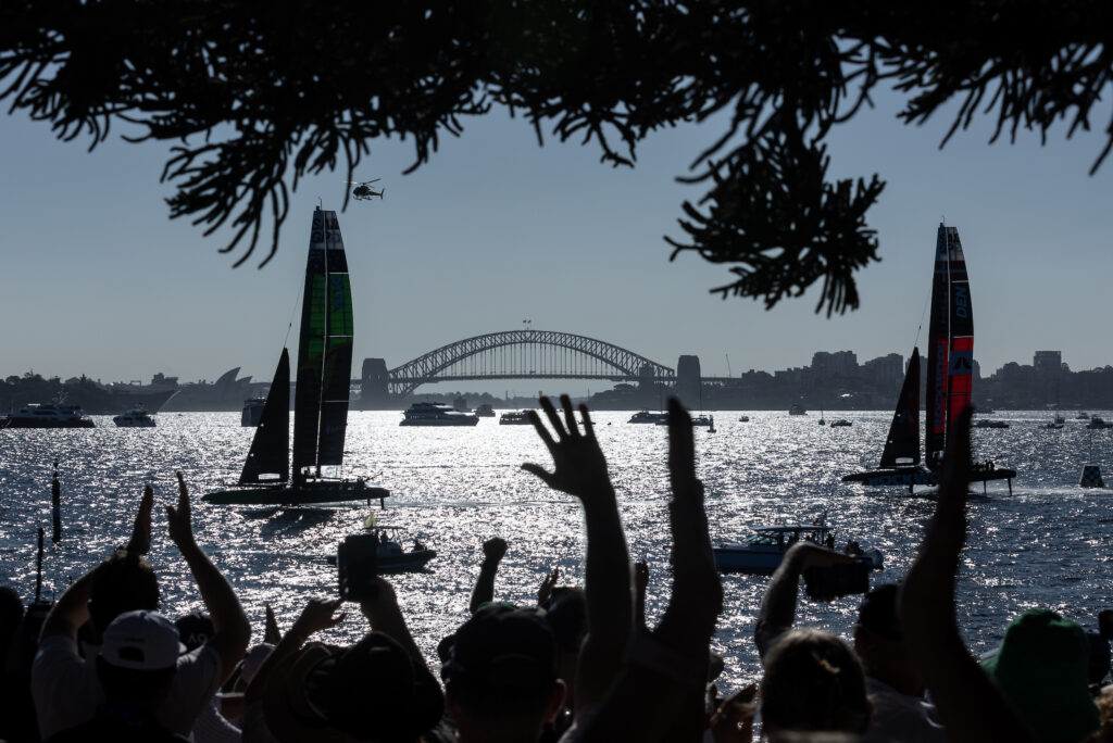 Silhouette of guests cheering as Australia SailGP Team and ROCKWOOL Denmark SailGP Team sail past Genesis Island with Sydney Harbour Bridge in the distance on Race Day 2 of the KPMG Australia Sail Grand Prix in Sydney, Australia. Saturday 25th February 2024. Photo: Patrick Hamilton for SailGP. Handout image supplied by SailGP