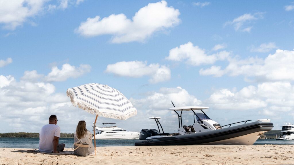 People under parasol on beach looking at Grand Boats vessel in the sea.