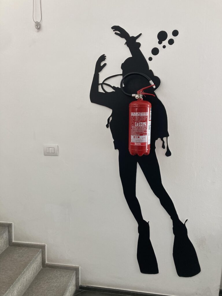 fire extinguisher on wall with diver painted behind it so extinguisher looks like oxygen bottle - at Osculati
