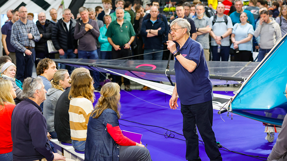 A male delivers a talk to a crowd who sit and stand around him at the RYA Dinghy and Watersports Show