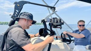 One man carrying a camera films another man - who is driving a boat.
