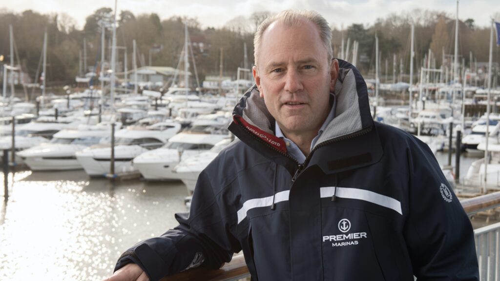 Man in jacket smiles at camera as he stands in front of marina full of boats