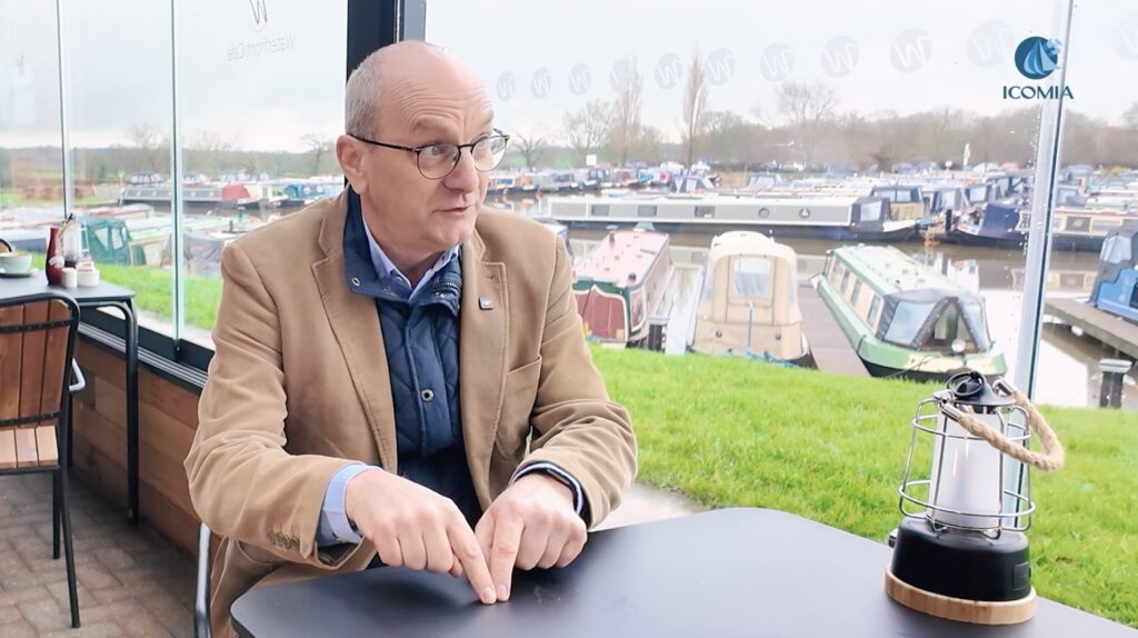 Man in beige jacket sits at table talking and out the window behind is canal boats