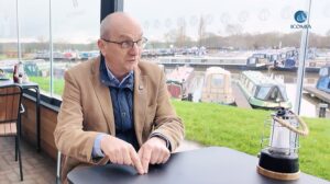 Man in beige jacket sits at table talking and out the window behind is canal boats