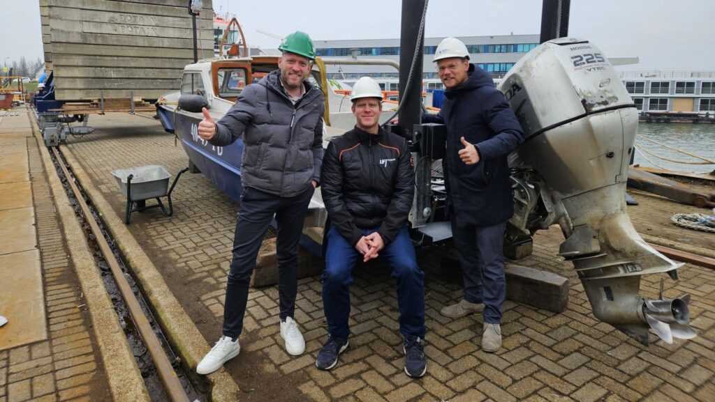 Three men lean on a boat and - wearing hard hats - make thumbs-up signs
