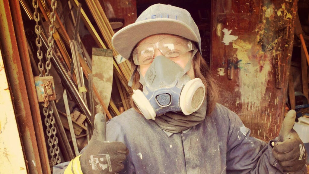 boatbuilder in overalls and assorted PPE gives a dusty thumbs-up to the camera