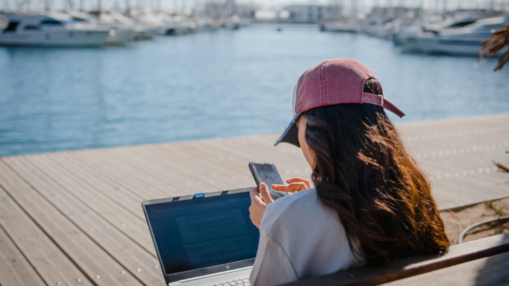 A woman sitting on a bench at a marina, using a laptop computer