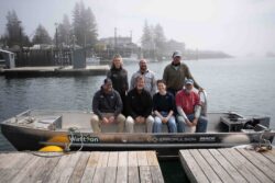 7 people standing and sitting on a skiff boat on a lake