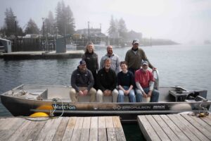 7 people standing and sitting on a skiff boat on a lake
