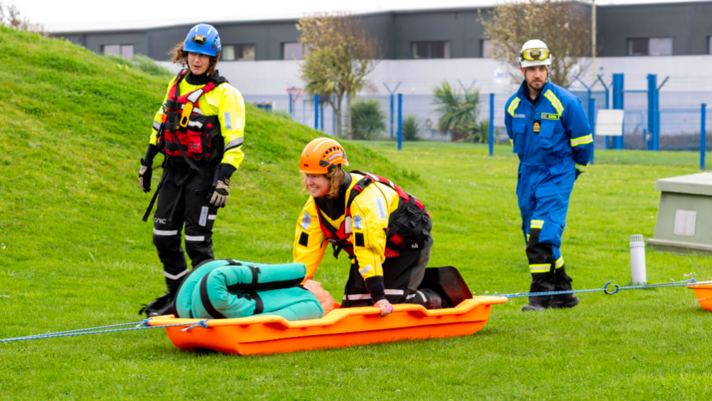 three people in protective gear and a sledge on grass - part of a training excercise