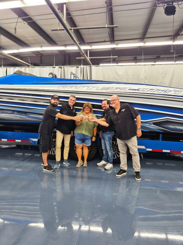Five people raise glasses in front of an Outerlimits performance boat inside the company’s factory