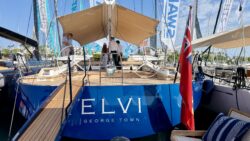 A blue sailing yacht named ELVI moored at a marina, with a large beige cockpit awning and matching upholstered seating.