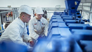 Two trainees wearing hard hats and protective clothing inspect industrial machinery in a workshop.